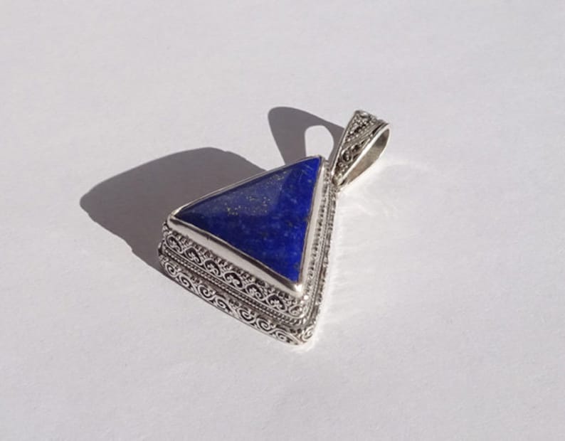 May include: A triangular pendant with a deep blue stone set in an ornate silver-colored metal frame. The pendant has a decorative bail for a chain. The design features detailed filigree work around the stone.