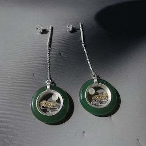 Peut inclure: Une paire de boucles d'oreilles pendantes en argent avec des cercles en jade vert. Les cercles de jade ont un cadre en argent avec un motif de lune et de nuages dorés à l'intérieur.