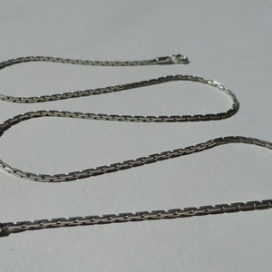 Peut inclure: Un collier en chaîne argent avec un design de chaîne à maillons carrés. La chaîne mesure environ 50 cm de long et se ferme avec un fermoir à mousqueton.