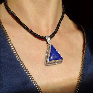 May include: A silver pendant with a triangular, deep blue stone hangs from a black cord necklace. The pendant is framed with a detailed silver border. The necklace is worn against a dark blue garment.