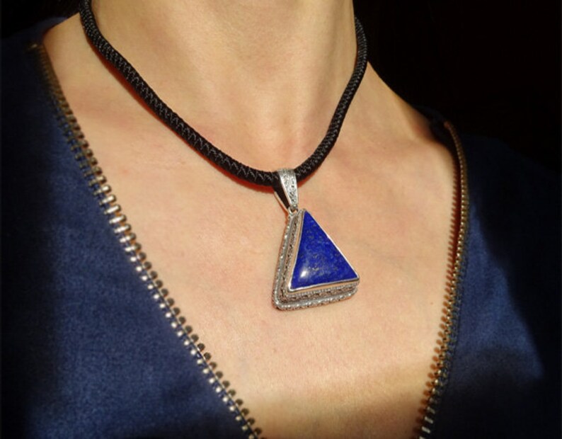 May include: A silver pendant with a triangular, deep blue stone hangs from a black cord necklace. The pendant is framed with a detailed silver border. The necklace is worn against a dark blue garment.