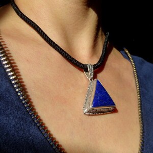 May include: A triangular pendant necklace with a deep blue stone set in a silver-colored metal frame. The pendant hangs from a black cord necklace. The person is wearing a blue top with a zipper.