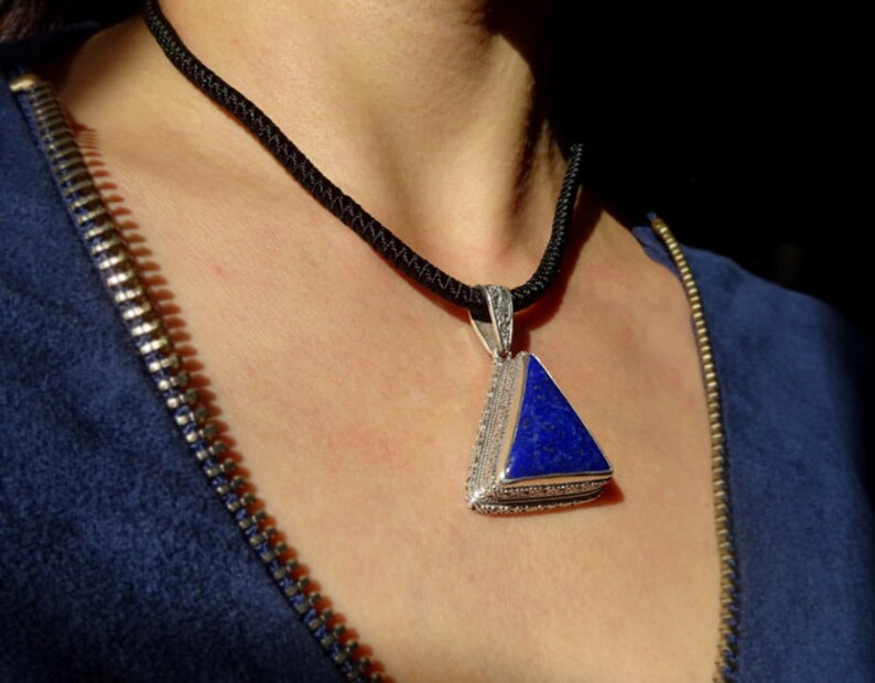 May include: A triangular pendant necklace with a deep blue stone set in a silver-colored metal frame. The pendant hangs from a black cord necklace. The person is wearing a blue top with a zipper.
