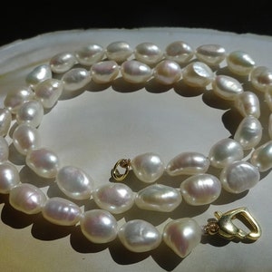 Può includere: Una collana di perle bianche con una chiusura dorata. Le perle sono di forma ovale e hanno una leggera lucentezza iridescente.
