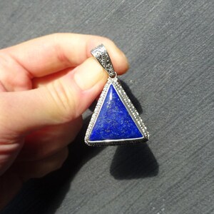 May include: A triangular pendant with a deep blue stone set in a silver-colored metal frame. The stone has a speckled appearance, and the frame is detailed with small, decorative elements. The pendant is suspended from a matching bail.
