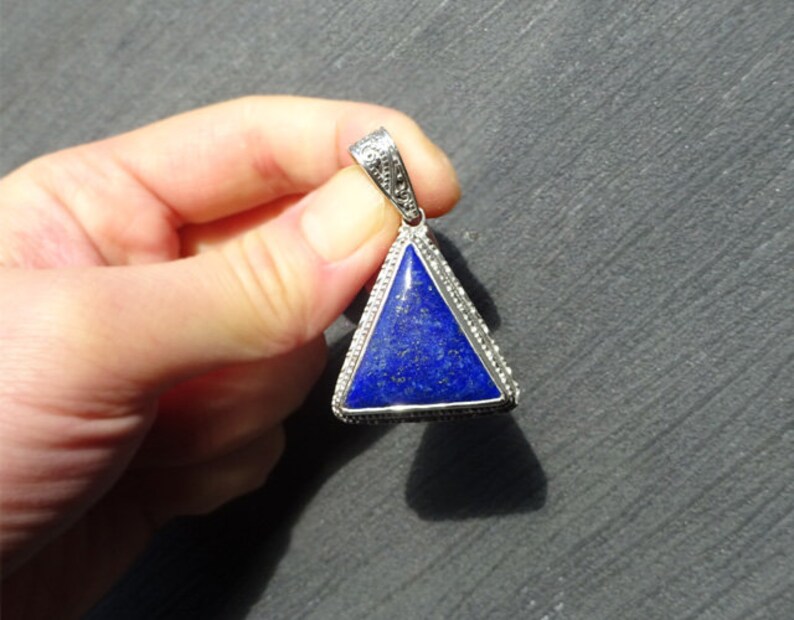 May include: A triangular pendant with a deep blue stone set in a silver-colored metal frame. The stone has a speckled appearance, and the frame is detailed with small, decorative elements. The pendant is suspended from a matching bail.