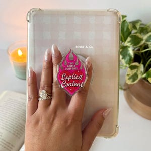 May include: Clear phone case with a pink and red graphic that reads "Explicit Content". The case is on a tablet. The brand name "Birdie & Co." is printed on the case. A hand with a ring and bracelet is holding the tablet.