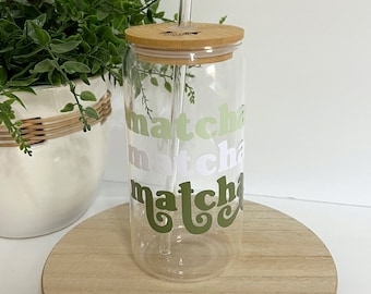 May include: A clear glass tumbler with a bamboo lid and a glass straw. The tumbler has the word "matcha" written on it in green, white, and pink lettering.