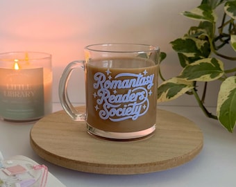 May include: A clear glass mug filled with a coffee-like beverage, featuring the text "Romantasy Readers Society" in a light blue font. The mug sits on a wooden coaster, with a lit candle and a book in the background.
