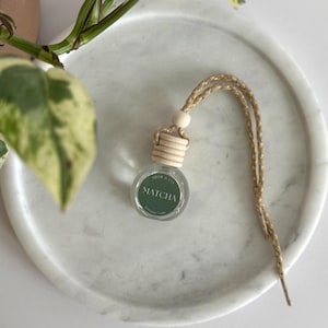 May include: A small, clear glass bottle with a wooden cap and a woven cord, labeled "Matcha" in green lettering. The bottle is on a white marble tray, with a green and white plant in the background. The cord is beige.