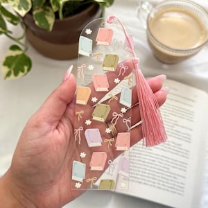 May include: Clear acrylic bookmark featuring a repeating pattern of colorful books, bows, and flowers. A pink tassel hangs from the top. The bookmark is held over an open book, with a cup of coffee and a potted plant in the background.