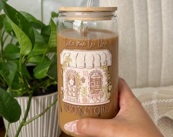 May include: A clear glass tumbler with a bamboo lid and a straw. The tumbler has a brown liquid inside and a graphic of a bookstore with the text "Take me to the bookstore".
