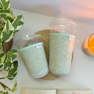 May include: Two clear plastic cups with lids, each encased in a light green floral patterned sleeve. The sleeves have a mint green trim and a tag that reads "MADE & CO." The cups sit on a wooden coaster, next to a lit candle and a houseplant.