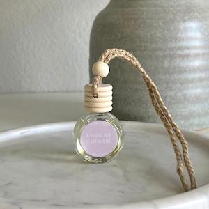 May include: A small, clear glass bottle with a wooden cap and a pink label that reads "Lavender Cashmere". The bottle is hanging from a brown string and is sitting on a white marble surface.