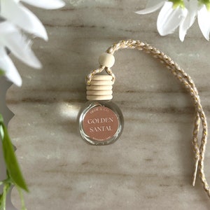 May include: A small glass bottle with a wooden cap and a gold string. The bottle is filled with a clear liquid and has a label that reads "GOLDEN SANTAL" with the brand name "FRED & CO."
