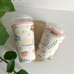May include: Two reusable coffee cup sleeves with a pink trim. The sleeves feature a light blue background with a pattern of books, flowers, and teacups.