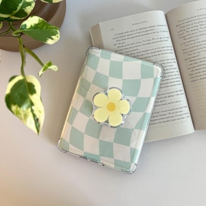 May include: A clear phone case with a light blue and white checkered pattern. A yellow flower-shaped phone grip is attached. The case is resting on a book with visible text and a green plant.