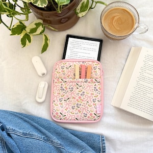 May include: A pink floral e-reader case with a matching pink trim, holding an e-reader with text. The case has a front pocket with three coloured page markers. A cup of coffee, a book, and a denim jacket are also in the image.