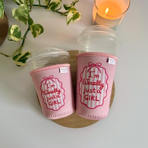 May include: Two pink drink sleeves with the text "I'm literally just a GIRL" in a white and pink striped design. The sleeves are on clear plastic cups with lids. A lit candle and greenery are in the background.