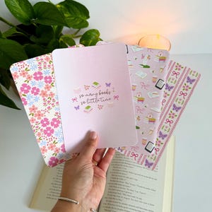 May include: A set of four rectangular bookmarks with varied designs including a pink floral pattern, a solid light pink card with the text so many books so little time, a light purple pattern with books and coffee, and a pink ribbon and butterfly print.