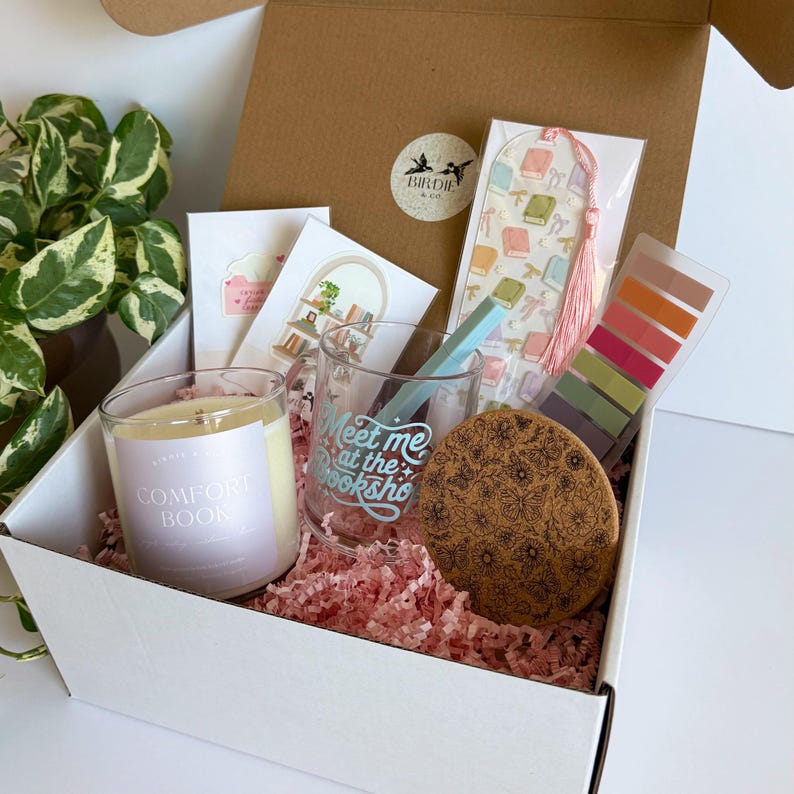 May include: A gift box filled with a candle, a glass with the text "Meet me at the bookshop", a bookmark, sticky notes, a coaster, and cards. The box is lined with pink paper and includes a plant.