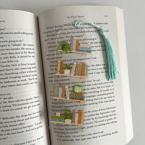 Pu&ograve; includere: Un primo piano di un libro aperto con un segnalibro. Il segnalibro &egrave; un fiocco blu chiaro con una pila di libri colorati e una piccola pianta in vaso. Le pagine del libro sono piene di testo e il segnalibro copre parzialmente il testo. Il testo sulla pagina recita: "Non &egrave;", dice. "Hai ragione per quanto riguarda la tua et&agrave;. Conta molto. Combatter&ograve; per te, ma ho bisogno di te qui con me."