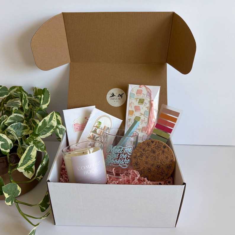 May include: Open gift box filled with a candle, bookmark, glass, cards, and a coaster. The candle reads "Comfort Book". The glass reads "Meet me at the Booksho". The box is lined with pink paper.
