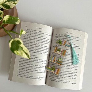 Pu&ograve; includere: Primo piano di un libro con un segnalibro che raffigura una pila di libri e una pianta in vaso. Il segnalibro &egrave; in plastica trasparente e ha un fiocco blu chiaro. Il testo sul libro dice "In Five Years".