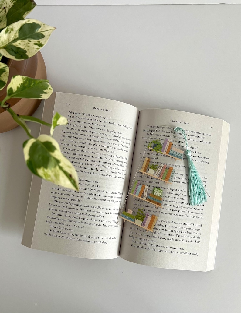 Pu&ograve; includere: Primo piano di un libro con un segnalibro che raffigura una pila di libri e una pianta in vaso. Il segnalibro &egrave; in plastica trasparente e ha un fiocco blu chiaro. Il testo sul libro dice "In Five Years".