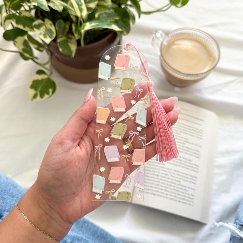 May include: Clear acrylic bookmark featuring a repeating pattern of colorful books, bows, and flowers. A pink tassel hangs from the top. The bookmark is held over an open book, with a cup of coffee and a potted plant in the background.