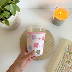 May include: A pink coffee cup sleeve with a pattern of colorful books and bows. The sleeve is wrapped around a clear plastic cup with a lid. A lit candle and a book are in the background.