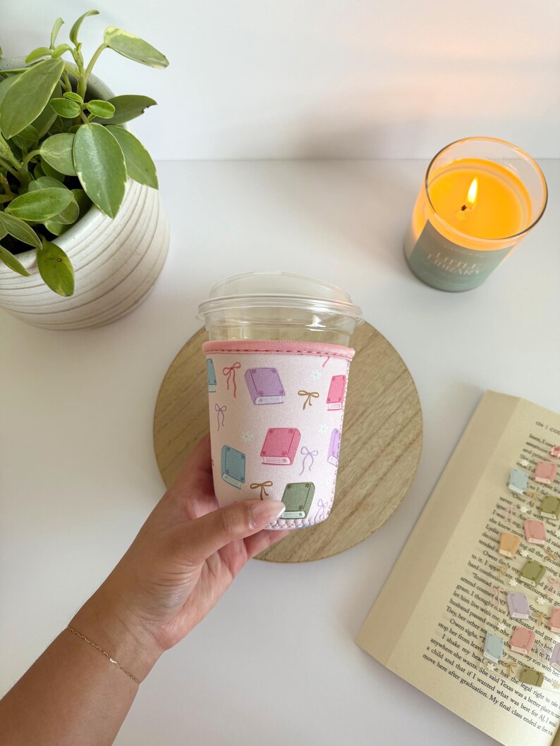 May include: A pink coffee cup sleeve with a pattern of colorful books and bows. The sleeve is wrapped around a clear plastic cup with a lid. A lit candle and a book are in the background.