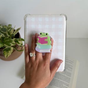 May include: A clear tablet case with a light pink and white pattern. A clear acrylic phone grip featuring a green frog reading a pink book is attached to the case. The phone grip is on top of a stack of books. The text "Birdie & Co" is visible.