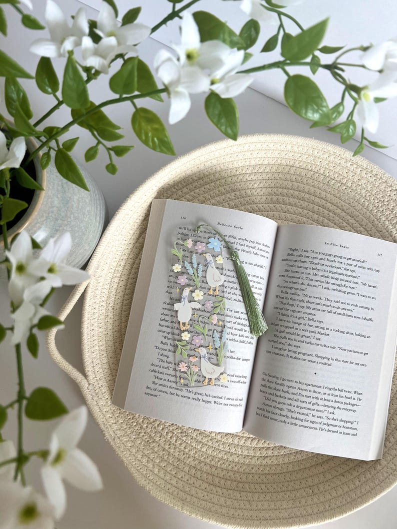 May include: A close-up of a book with a bookmark featuring a floral design and a group of ducks. The bookmark has a green tassel. The book is open to a page with the text "In Five Years" by Rebecca Serle.