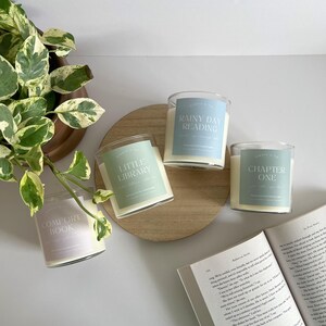 May include: Four glass jar candles with labels that read "Comfort Book", "Little Library", "Rainy Day Reading", and "Chapter One". The candles are on a wooden tray with a book and a plant in the background.