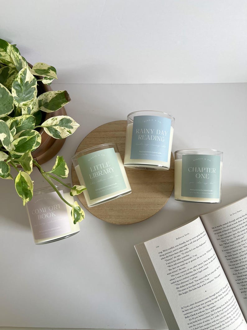 May include: Four glass jar candles with labels that read "Comfort Book", "Little Library", "Rainy Day Reading", and "Chapter One". The candles are on a wooden tray with a book and a plant in the background.