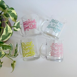 May include: Four clear glass mugs with curved handles. Each mug has a different phrase in colorful text: "Antisocial Book Club", "Meet me at the Bookshop", "Read more Books", and "I Read Past my Bedtime". A green and white plant is in the background.