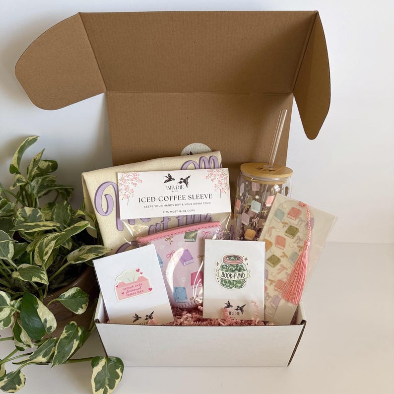 May include: An open cardboard box filled with a variety of gifts, including an iced coffee sleeve, a glass tumbler, a bookmark with a pink tassel, and small cards. The box is lined with pink crinkle paper and includes a potted plant.