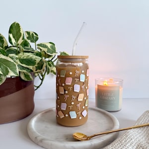 May include: A clear glass tumbler with a wooden lid and a clear straw, filled with a brown beverage. The tumbler is decorated with pastel-colored books and bows. It sits on a marble tray with a gold spoon, next to a lit candle and a plant.