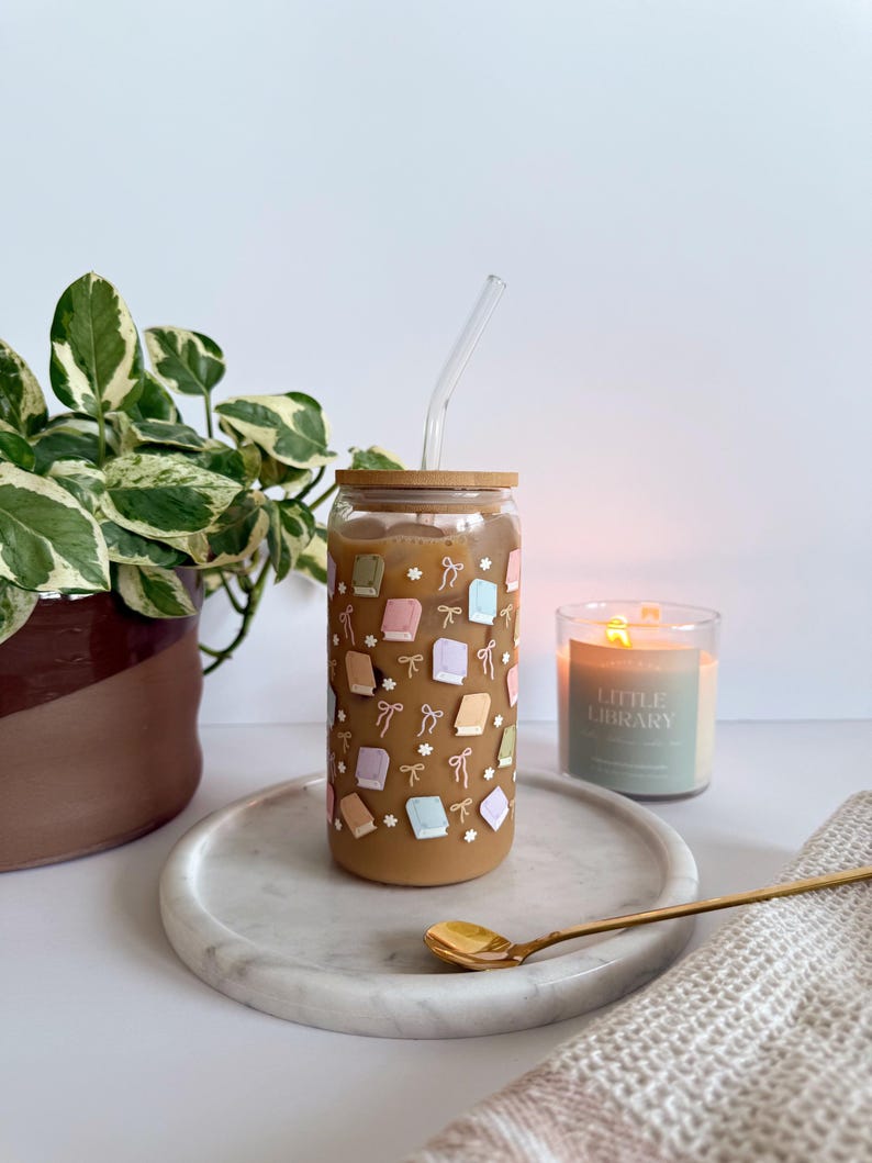 May include: A clear glass tumbler with a wooden lid and a clear straw, filled with a brown beverage. The tumbler is decorated with pastel-colored books and bows. It sits on a marble tray with a gold spoon, next to a lit candle and a plant.