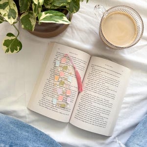 May include: An open book with a colorful bookmark featuring miniature book charms and a pink tassel. A cup of coffee sits nearby, with a potted plant in the background. The bookmark is made of clear beads and small, pastel-colored book-shaped charms.