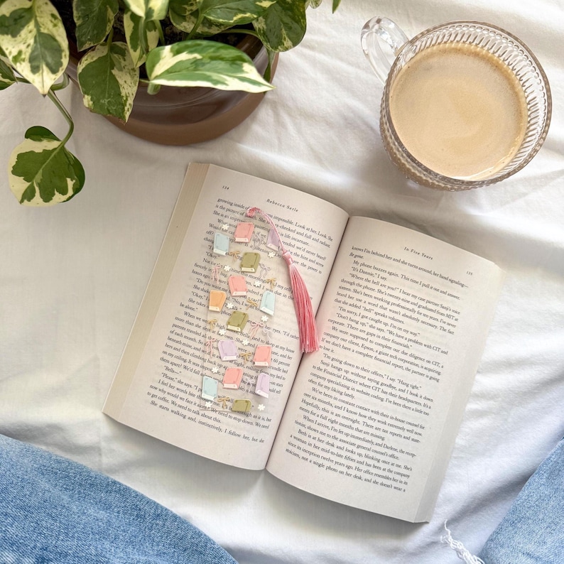 May include: An open book with a colorful bookmark featuring miniature book charms and a pink tassel. A cup of coffee sits nearby, with a potted plant in the background. The bookmark is made of clear beads and small, pastel-colored book-shaped charms.