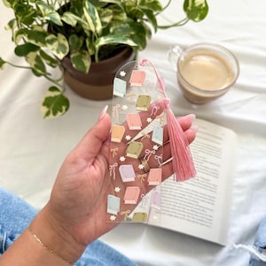 May include: Clear acrylic bookmark featuring pastel-colored book illustrations and bows. A pink tassel hangs from the top. The bookmark is held over an open book, with a cup of coffee and a potted plant in the background.