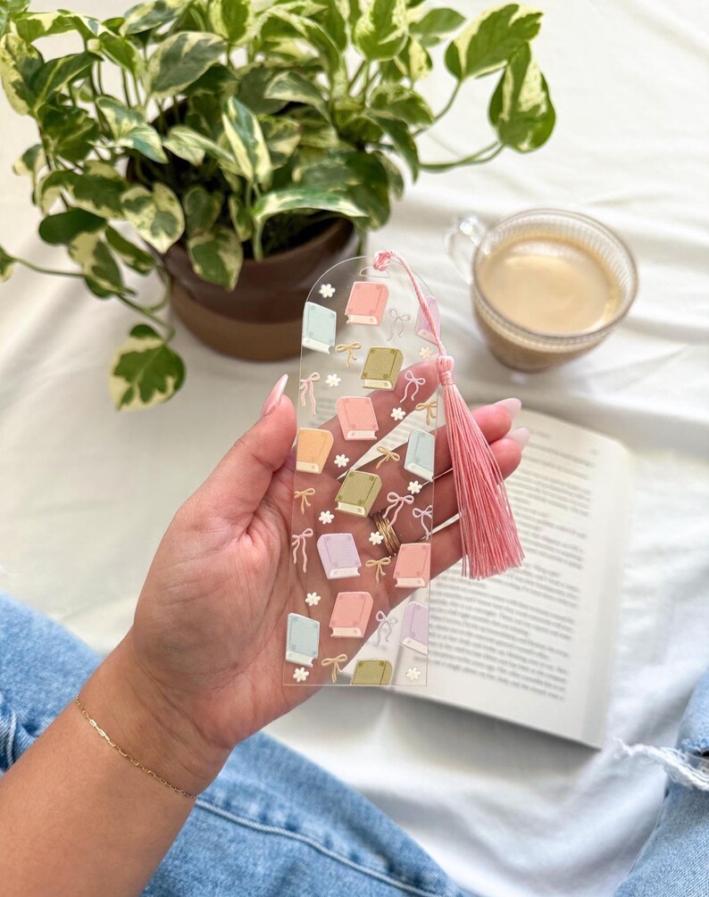 May include: Clear acrylic bookmark featuring pastel-colored book illustrations and bows. A pink tassel hangs from the top. The bookmark is held over an open book, with a cup of coffee and a potted plant in the background.