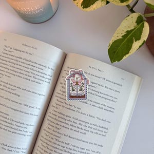 May include: A decorative bookmark with a whimsical design of an open book, flowers, and a dragonfly within a framed border. The bookmark is on a book, with a candle and plant in the background.