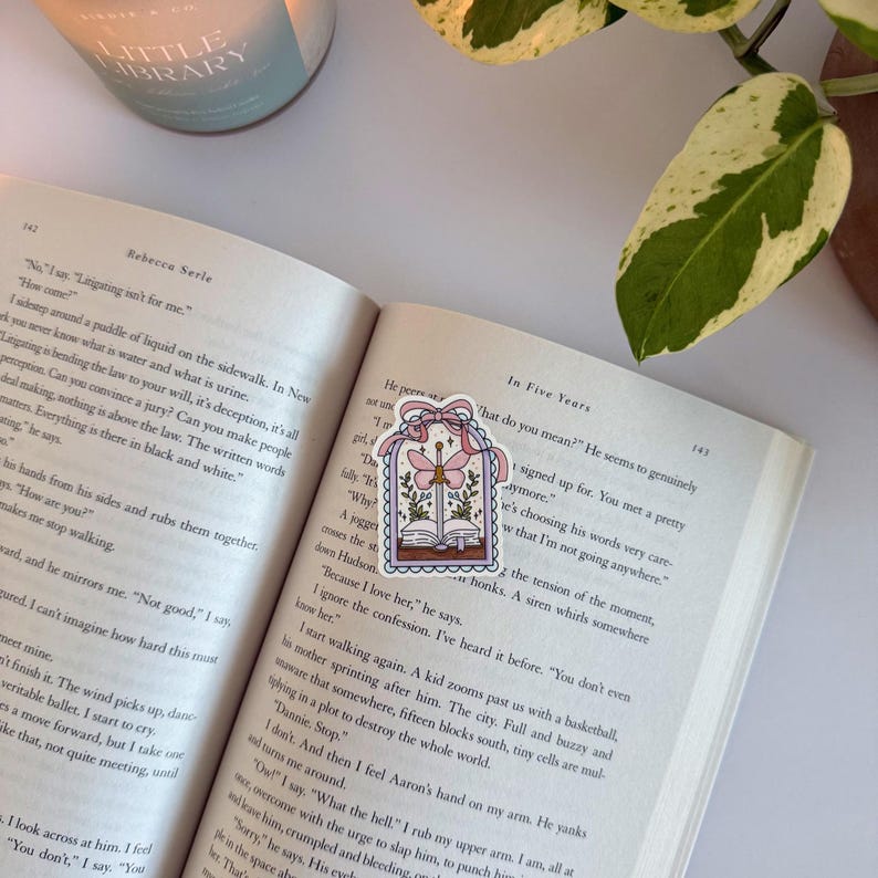 May include: A decorative bookmark with a whimsical design of an open book, flowers, and a dragonfly within a framed border. The bookmark is on a book, with a candle and plant in the background.