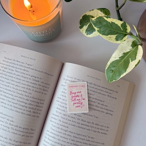 May include: A lit candle in a glass jar next to an open book with a pink library card that reads "Buy me books & Tell me I'm pretty" with hearts. A green and white plant is in the background.