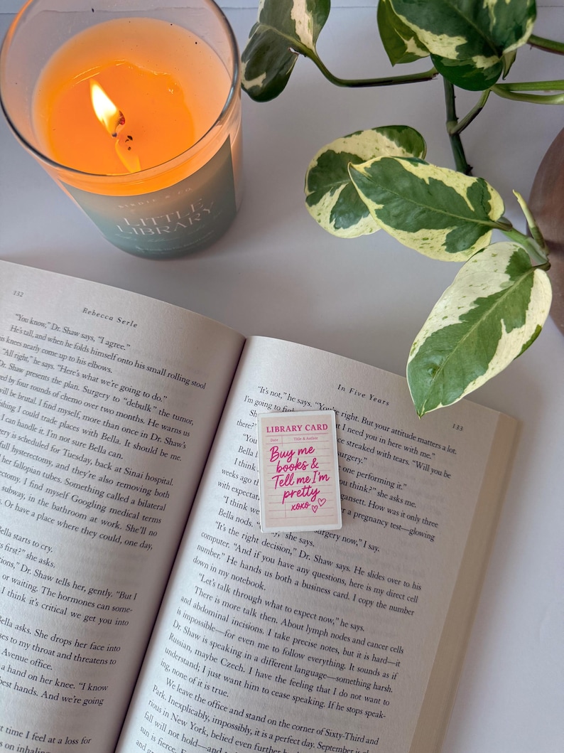 May include: A lit candle in a glass jar next to an open book with a pink library card that reads "Buy me books & Tell me I'm pretty" with hearts. A green and white plant is in the background.