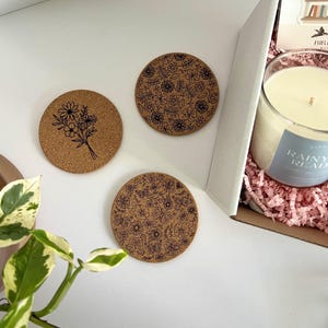May include: Three round cork coasters with black floral designs. One coaster features a bouquet, while the others display intricate patterns of flowers and leaves. A candle in a glass jar with a blue label that reads "Rainy Read" is visible in a box.