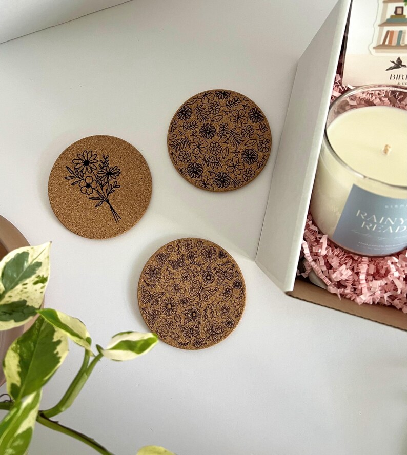 May include: Three round cork coasters with black floral designs. One coaster features a bouquet, while the others display intricate patterns of flowers and leaves. A candle in a glass jar with a blue label that reads "Rainy Read" is visible in a box.